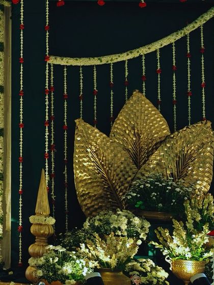 An elegant decor piece featuring a traditional throne against a dark green backdrop, accented with golden palm leaves and strings of jasmine flowers.