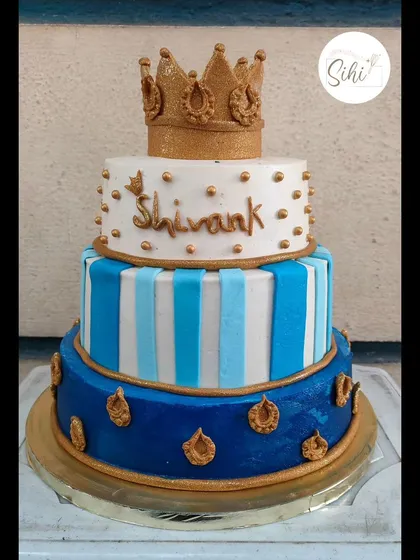 A royal three-tier cake fit for a little prince. This design features a bold blue and white color scheme with stripes, gold accents, and a glittering gold crown topper.