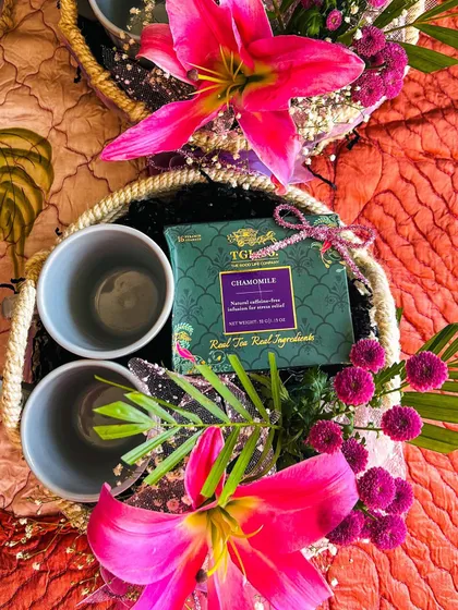 A vibrant floral hamper designed as a wedding or corporate favour. This basket features two ceramic mugs and a box of chamomile tea, surrounded by bright pink lilies and other colourful flowers, making it a refreshing and beautiful gift.