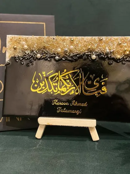 This personalized plaque features the verse "Fabi ayyi ala i rabbikuma tukazziban" in bold yellow calligraphy on a black background. The top is adorned with a textured mix of gold and silver embellishments, and it includes the client's name for a personal touch.