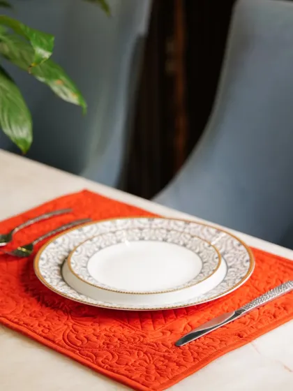 A bright red quilted placemat provides a pop of color on a dining table. The simple, clean setup with elegant dinnerware highlights the placemat's texture and vibrant hue.
