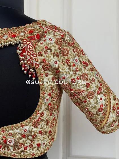 The back of the off-white wedding procession blouse. It features a deep, wide cutout with a pearl tassel detail at the top, framed by the same intricate floral and traditional motif embroidery.