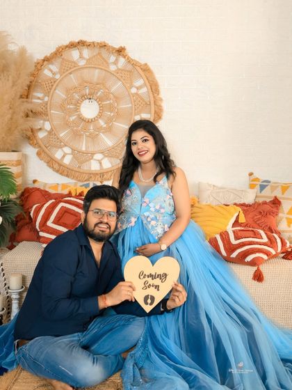 A happy couple portrait in our cozy, boho-themed setup. They hold a "Coming Soon" sign, sharing a joyful moment surrounded by textured pillows and pampas grass.