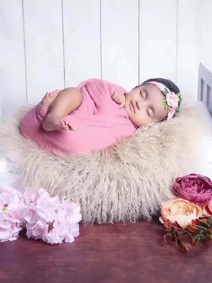 A sleeping baby girl in a pink wrap and floral headband, posed in a miniature white bed against a simple, clean background.