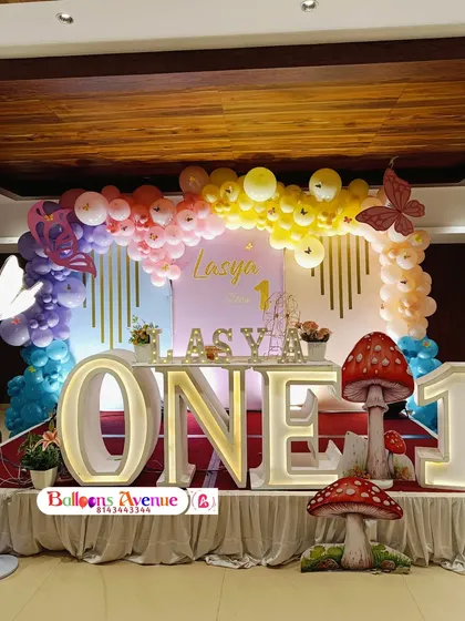 A vibrant rainbow butterfly theme for Lasya's first birthday. This stage setup includes a large 'ONE' table, mushroom props, and a colorful balloon arch.