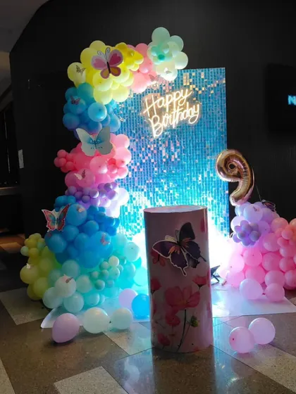 A colorful butterfly theme decoration against a blue sequin wall. The setup includes a rainbow-colored balloon column and a custom-printed pedestal, perfect for a vibrant birthday celebration.