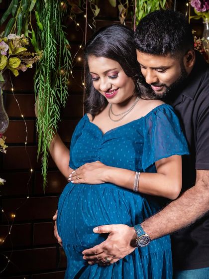 A tender moment in a rustic setting. The partner embraces the mom-to-be from behind, both looking down at the bump with love, surrounded by warm fairy lights.