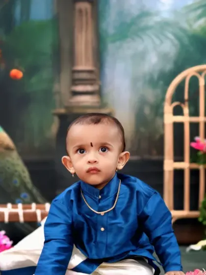 A toddler in a traditional blue kurta and veshti, posing sweetly in a setting decorated with flowers and a peacock.