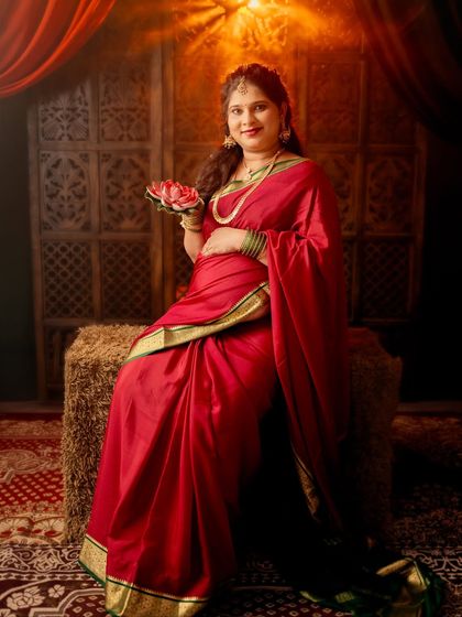 A regal portrait of a mom-to-be seated on a rustic stool. The rich red saree and traditional props create a feeling of timeless elegance.