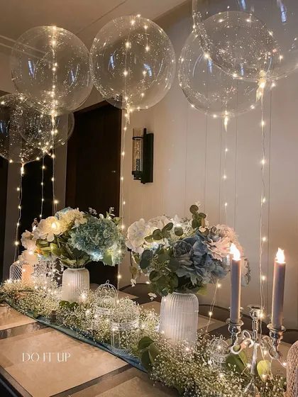 A magical indoor dinner setting. The table is decorated with a runner of baby's breath and hydrangeas, while fairy-light-filled balloons float above, creating an ethereal and dreamy atmosphere.