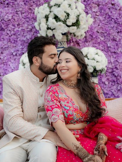 A tender moment where the groom kisses the bride's cheek. Their joyful expressions and the vibrant floral background make this a perfect Mehendi portrait.