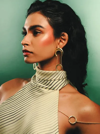 A portrait showcasing a different set of Batoki Art jewelry, including long, circular earrings. The striped top and green background provide a contrasting graphic element to the elegant gold pieces.