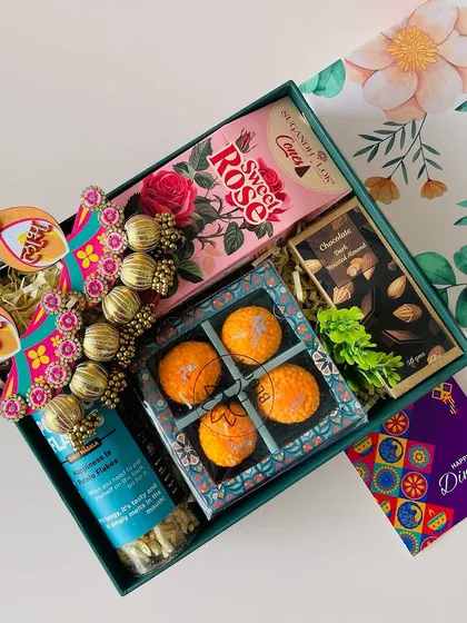 This floral box hamper offers a choice of contents. This version includes festive sweets, "Shubh Labh" hangings, a chocolate bar, and snacks.