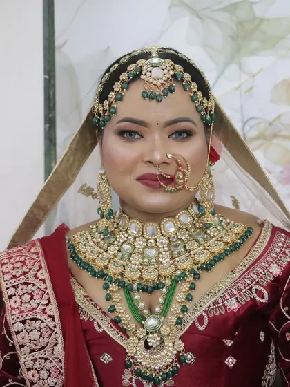A regal bridal look featuring a deep red lehenga and heavy Kundan jewellery with green accents. The makeup is bold and glamorous, with smokey eyes and a matte red lip to create a truly royal appearance.