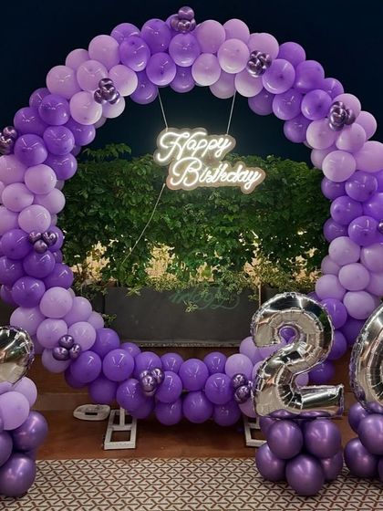 A beautiful 26th birthday decoration in shades of purple. The setup includes a full circular balloon arch, a 'Happy Birthday' neon sign, and silver foil number balloons.