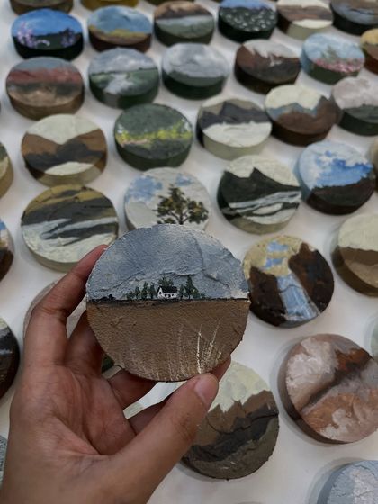 This miniature painting depicts a rustic house under a cloudy sky. Each piece is hand-painted on a textured MDF board.