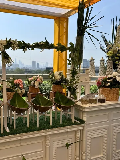 A fresh and airy outdoor buffet setup with a view of the city skyline. The white counters are adorned with green foliage and wicker baskets, creating a chic and natural vibe.