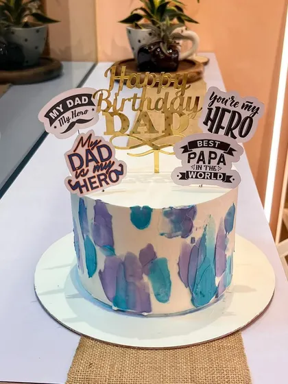 A cake to celebrate the hero of the family. This Father's Day cake has a cool blue and purple watercolor finish and is decorated with various "My Dad, My Hero" toppers.