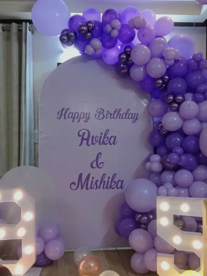 A beautiful purple-themed decoration for a joint birthday celebration. The custom arched backdrop is framed by a balloon garland in various shades of purple, complemented by large light-up numbers '9' and '5'.