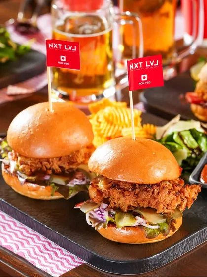 Two delicious-looking sliders with crispy chicken, served with chips and a side salad. The mugs of beer in the background firmly place this shot within an Oktoberfest or pub theme.