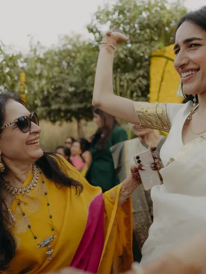 The mother and daughter dancing together at the Haldi. The mother's yellow and pink saree perfectly complements the festive decor and the bride's white outfit, creating a beautiful visual harmony.