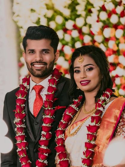 A smiling couple at their reception, framed by a wall of colorful flowers and their beautiful rose garlands.