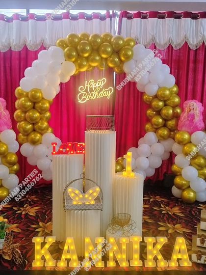 An elegant stage decoration with a gold and white balloon arch, Barbie props, and marquee letters.