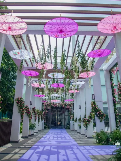 A whimsical and colorful walkway for a daytime engagement, featuring a purple carpet and a pergola ceiling decorated with hanging vines and Japanese-style umbrellas in shades of pink and white.