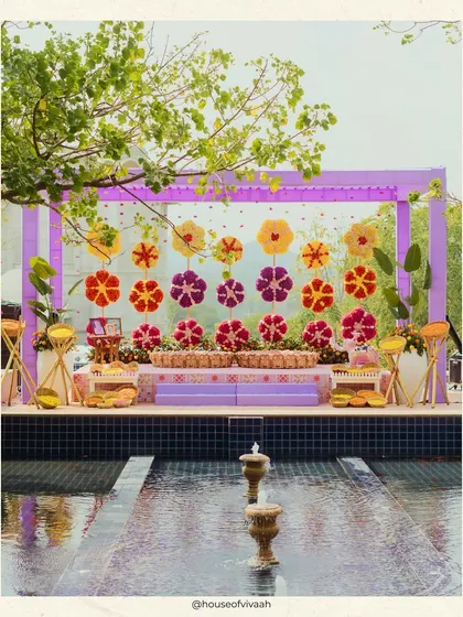 The Haldi stage set against a water feature, with a backdrop of giant floral lollipops and a canopy of lilac drapes, creating a stunning and unique ceremony space.