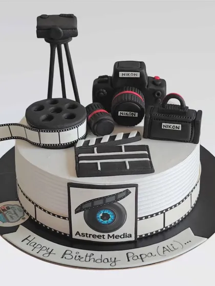 A detailed cake for a photographer and media professional. It features a fully edible Nikon camera, film reel, tripod, and clapperboard, all made from fondant.