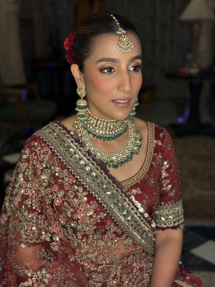 A portrait showcasing the complete bridal look. The focus is on flawless skin, softly defined eyes, and a neutral lip color that balances the richness of her red and gold embroidered outfit and heavy jewelry.