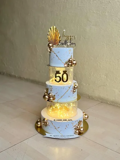 A grand 50th birthday cake with four tiers, using clear separators filled with fairy lights. The white and gold design is elegant and perfect for a milestone celebration.