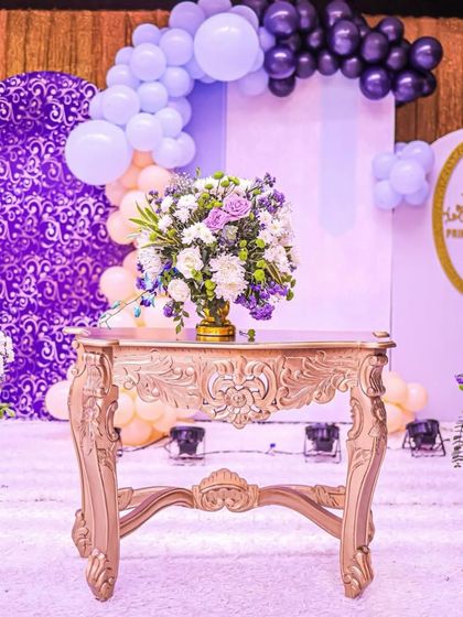 A close-up of the elegant floral arrangement and ornate table, with the purple and white balloon backdrop creating a soft, dreamy atmosphere for the royal celebration.