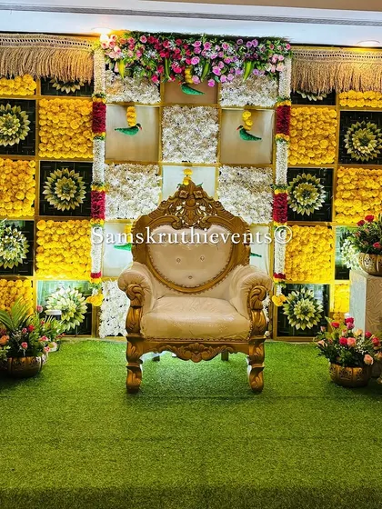 A classic baby shower setup with a checkerboard backdrop of yellow marigolds and woven green leaves. The traditional sofa and a full display of offerings on a striped mat complete this authentic South Indian look.