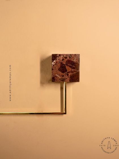 A close-up of a geometric wall sconce, featuring a square of rich, brown marble as its base. The combination of natural stone and a simple brass rod creates a fixture that is both minimalist and luxurious.