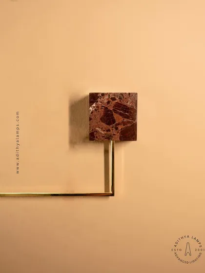 A close-up of a geometric wall sconce, featuring a square of rich, brown marble as its base. The combination of natural stone and a simple brass rod creates a fixture that is both minimalist and luxurious.