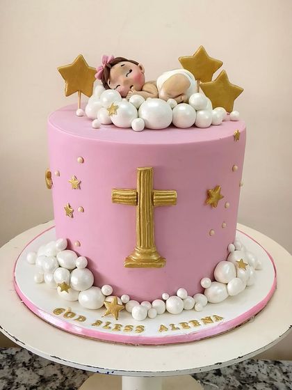 A dreamy christening cake in pink and gold. The design features a sleeping baby nestled in fluffy, pearlized clouds under golden stars, with a prominent cross on the front. It’s a sweet and serene cake for a blessed occasion.