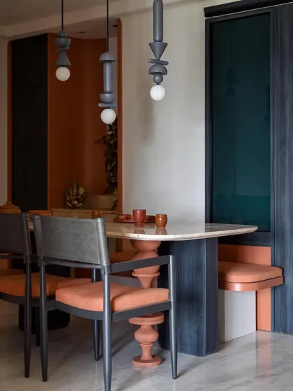 The dining area, with its travertine table, custom turned leg, and suspended wooden light fixtures. The minimalist mandir area in the background features a lotus symbol.