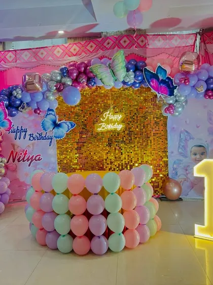 A beautiful butterfly-themed first birthday party. The decor features a gold shimmer wall, a pastel balloon arch with butterfly cutouts, and a marquee number '1'.