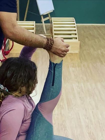An instructor provides hands-on assistance for a standing split against the wall, helping the student achieve greater height and better alignment.