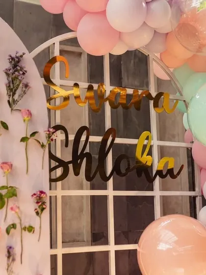 A custom gold acrylic name sign for Svara and Shloka's celebration. The backdrop includes a white grid frame and a pastel balloon arch, perfect for a baby shower or kids' party.