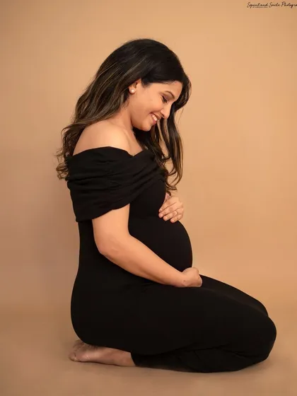 A serene and powerful solo maternity portrait. The mom-to-be kneels, cradling her belly in a classic black off-the-shoulder dress against a simple studio backdrop, highlighting the beauty of her pregnancy.