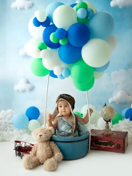A baby boy dressed as an aviator sits in a hot air balloon basket, surrounded by clouds and balloons. A creative and whimsical setup for a sitter session.