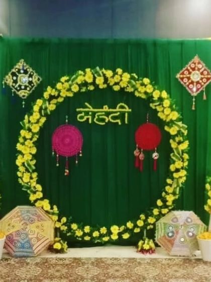 A simple and elegant Mehandi backdrop with a green drape, a marigold floral ring, and traditional hangings. The small, decorated Rajasthani umbrellas add a touch of folk art to the decor.
