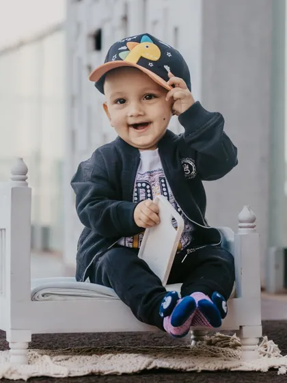 A cool little guy with a cap and a big smile, sitting on a miniature bed prop for his pre-birthday shoot.