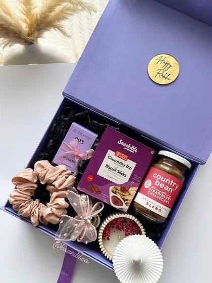 A lovely purple-themed Rakhi gift box for a sister, containing a scented candle, coffee, snacks, and scrunchies.