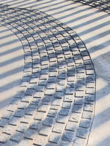 A close-up of the granite cobblestone paving, where the shadow patterns from the gate create a temporary, rhythmic inlay.