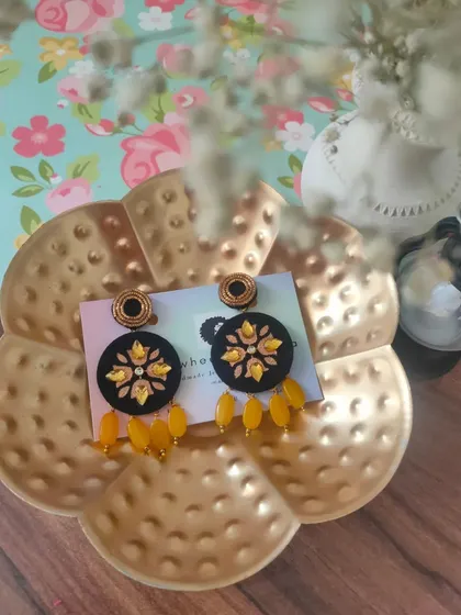 A pair of black fabric earrings with yellow floral embroidery and matching bead drops. The contrast of colors makes these a striking accessory for any occasion.