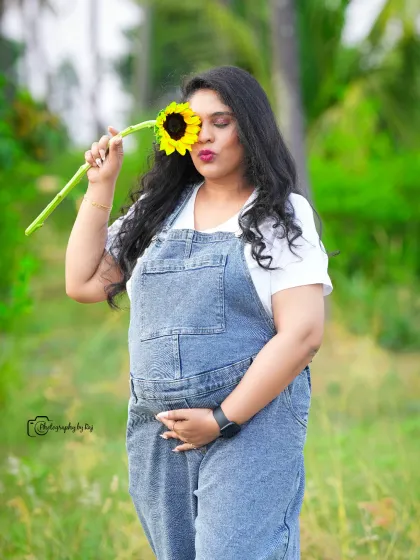 A playful solo portrait in denim overalls. The mother-to-be holds a sunflower over her eye, adding a touch of fun to this outdoor shot.