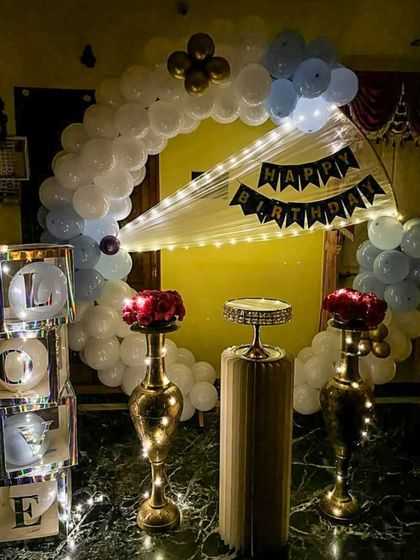 An elegant home setup featuring a white and light blue balloon arch. The decor includes tall brass vases with red roses, a central pedestal for the cake, and iridescent 'LOVE' blocks, creating a sophisticated party atmosphere.
